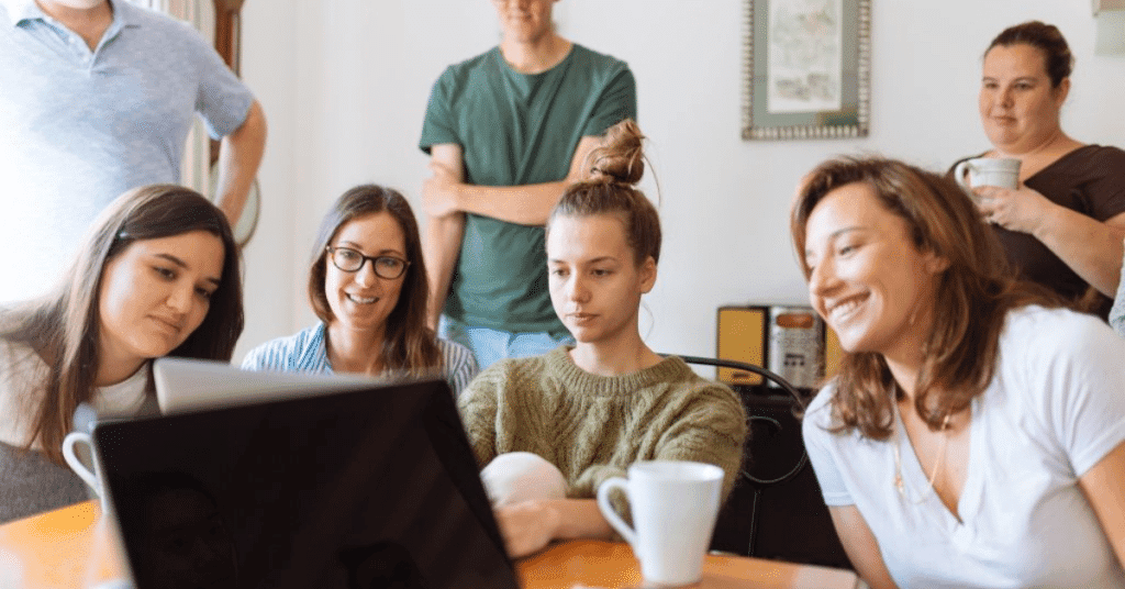 Four woman sitting and looking happy at a computer with some other colleagues behind them.