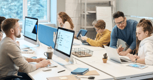 Multiple people from seemingly different ages sitting around a big table working, while one man is explaining something to a woman