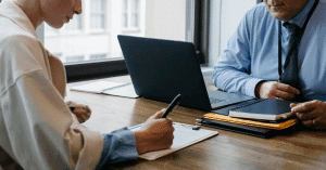 A woman writing and a men in front of a pc are sitting at a desk without seeing their faces.